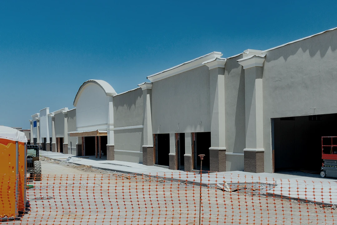 A large, unfinished commercial building with a plain facade is under construction behind orange safety netting, under a clear blue sky.