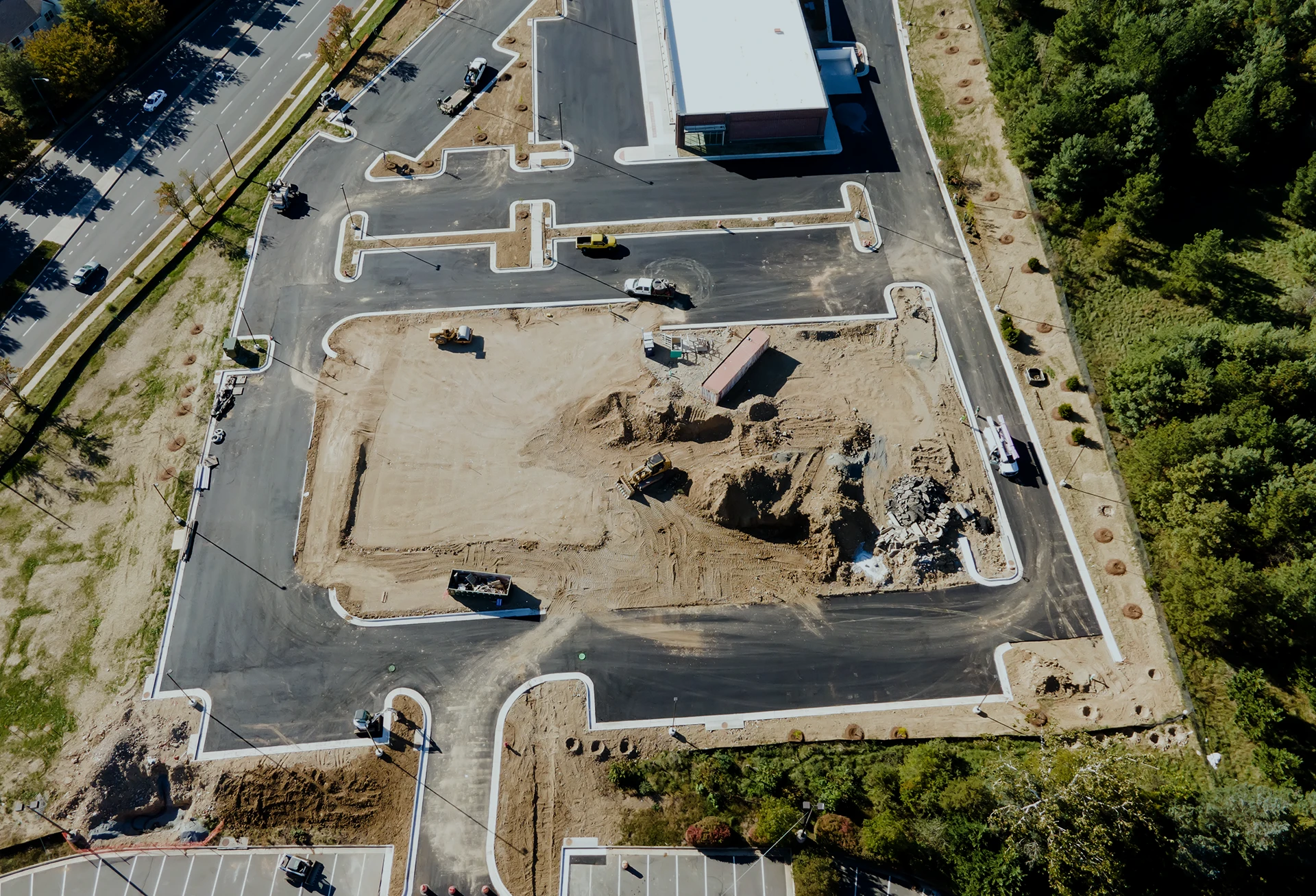 Aerial view of a construction site with dirt piles, machinery, and paved roads surrounding the area; nearby are trees and a parking lot.