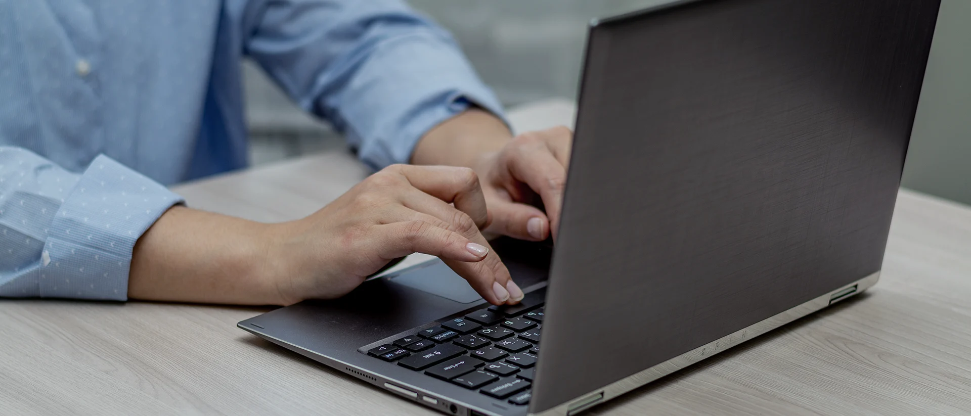 A person in a blue shirt is typing on a black laptop at a light-colored wooden desk. The image focuses on their hands and the laptop keyboard.