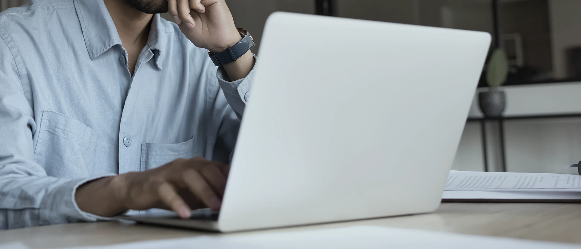 A person in a light blue shirt works on a white laptop at a desk, resting their chin on one hand while typing with the other. Papers and a pen are visible on the desk.