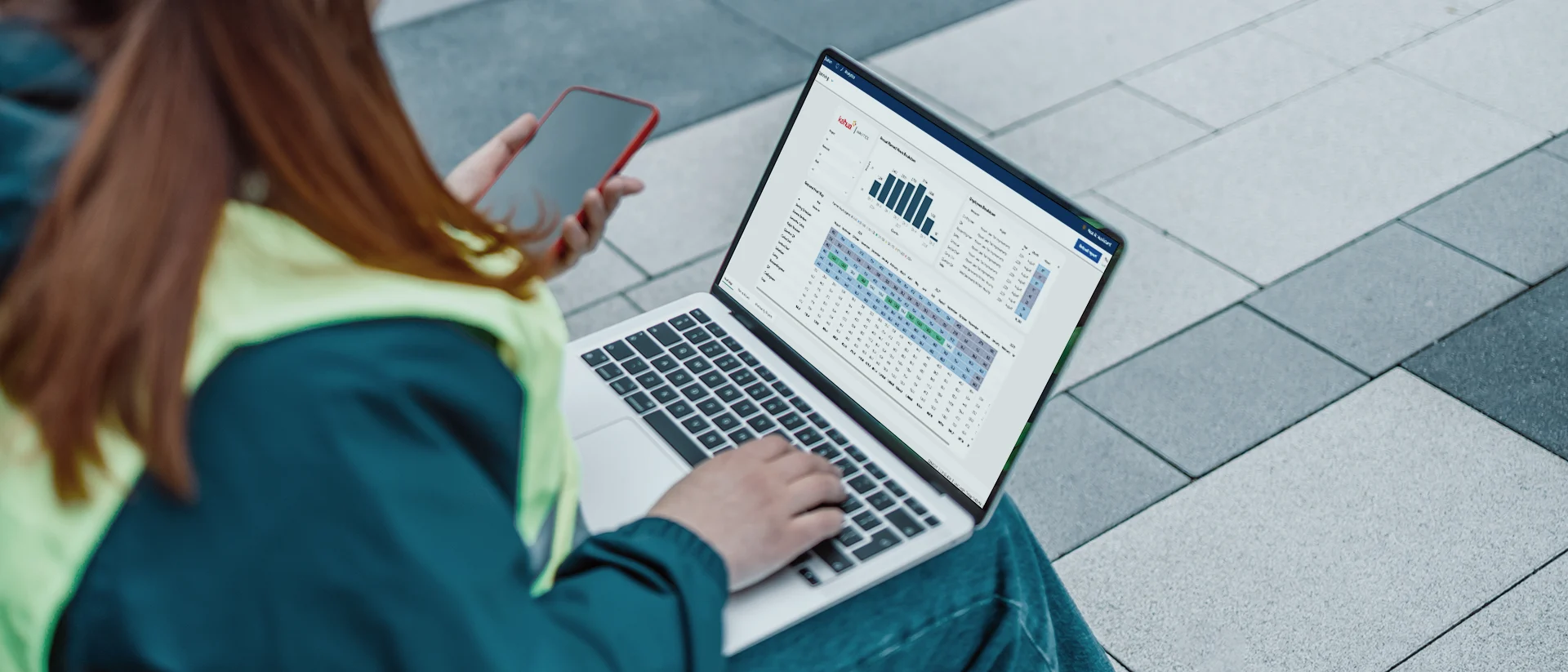 A person holding a phone and working on a laptop displaying data charts and graphs sits outdoors on gray tiled steps.