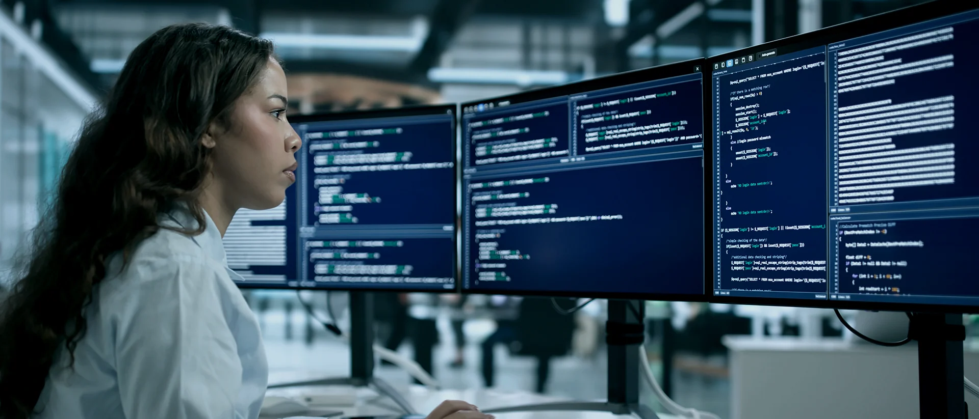 A woman sits at a desk in an office, focused on multiple large computer monitors displaying lines of code and technical information, suggesting she is working on software development or cybersecurity tasks.