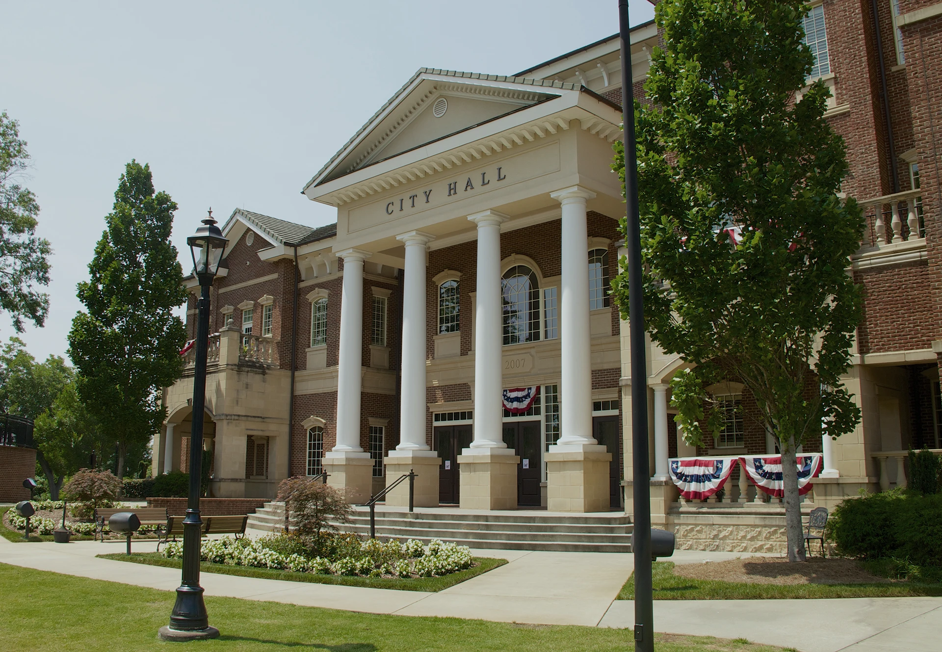 A brick city hall building with white columns, American flag bunting, and landscaped grounds with trees and flowers on a sunny day.