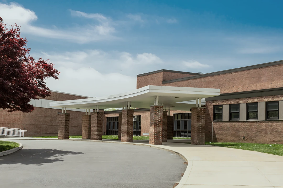 A modern brick school building with a covered entrance, surrounded by a driveway. The sky is blue with some clouds, and a tree with red leaves is visible on the left.