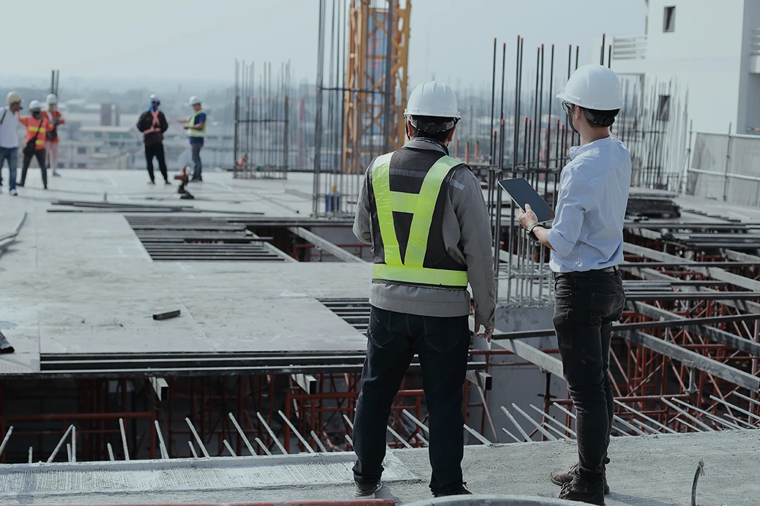 Two construction workers wearing safety gear and helmets stand on a building site, one holding a clipboard. In the background, other workers are visible near metal rods and scaffolding.