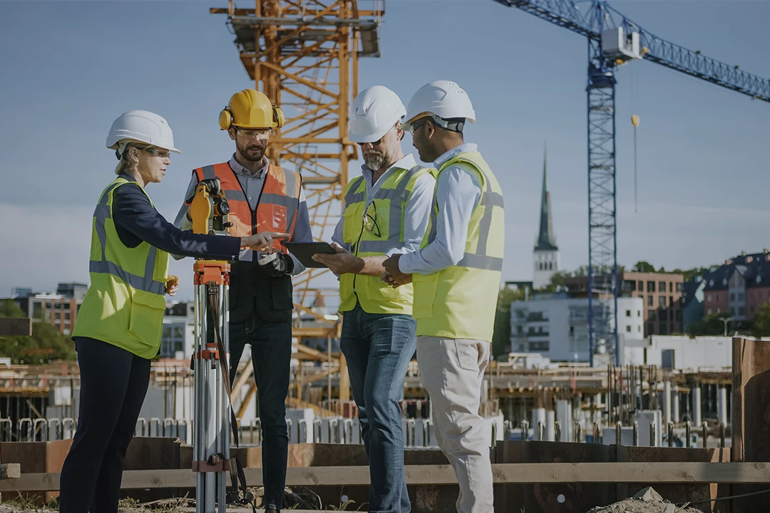 Four construction workers in safety gear discuss plans at a building site, with cranes and a cityscape in the background. One woman points at a tablet held by a man.