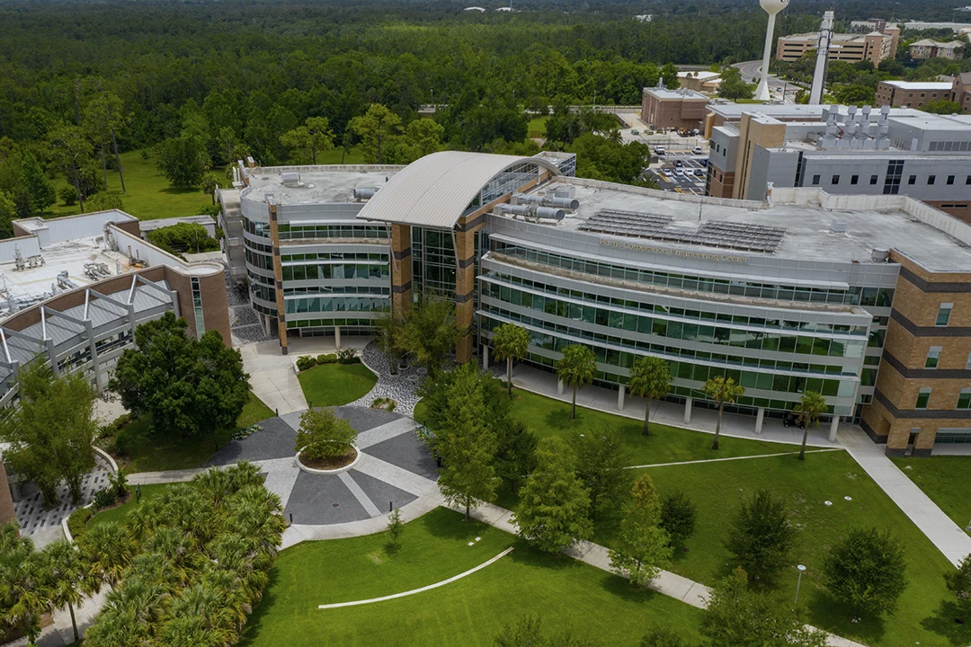 Aerial view of a modern, multi-story academic building with glass windows, curved architecture, and landscaped green lawns surrounded by trees.