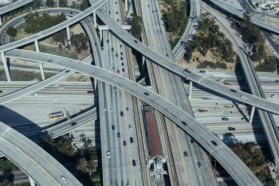 Aerial view of a complex highway interchange with multiple overpasses, underpasses, and cars traveling in different directions. Trees and greenery are visible alongside the roads.