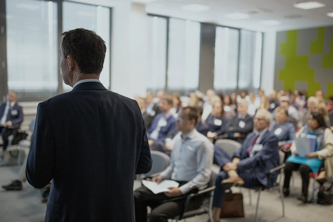 A man in a suit speaks to an audience seated in a modern conference room, with large windows and blurred attendees listening attentively.