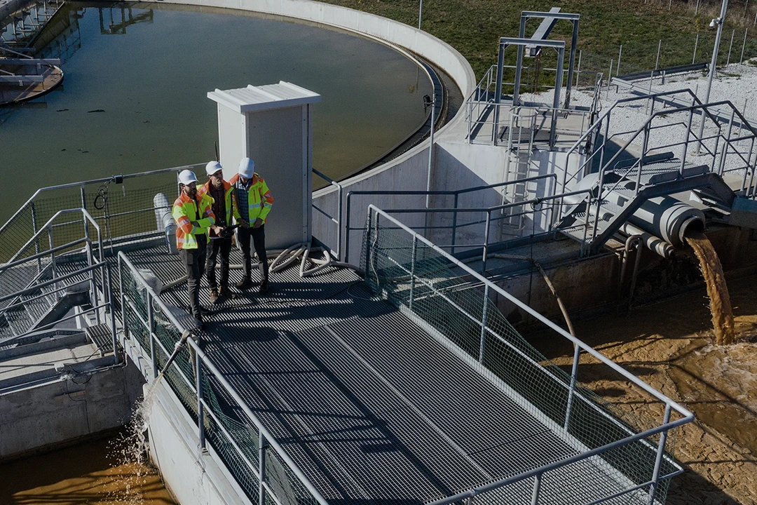 Three workers in safety vests and helmets stand on a metal walkway at a water treatment facility, inspecting the site as brown water flows through pipes into a basin.