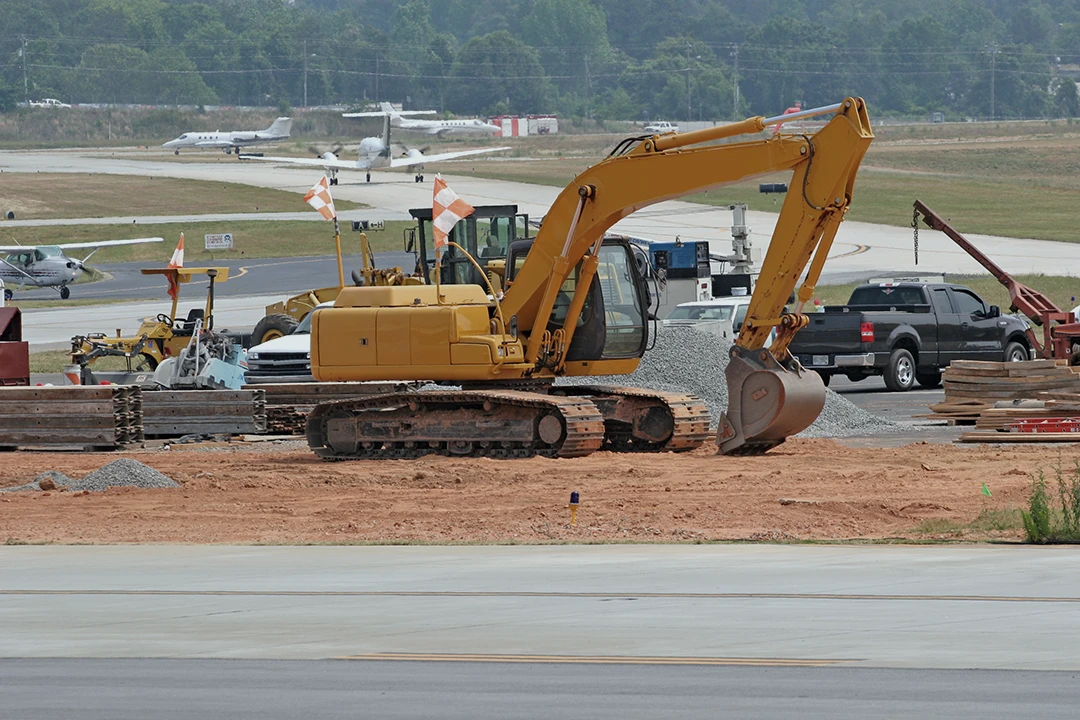 A yellow excavator is parked on a construction site near an airport runway, with piles of dirt, construction materials, vehicles, and small planes visible in the background.