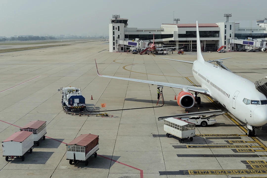 A white airplane is parked at an airport gate with ground service vehicles nearby. The terminal building and jet bridges are visible in the background on a clear day.