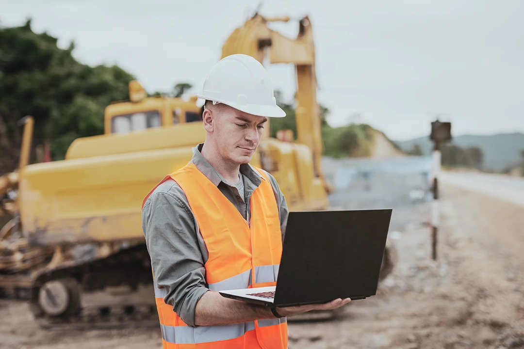 A construction worker in a white hard hat and orange safety vest uses a laptop at a construction site with heavy machinery in the background.