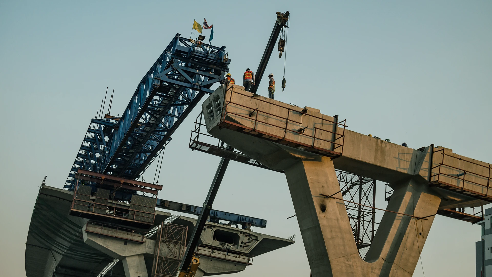 Construction workers stand atop a large concrete bridge segment, with cranes and scaffolding visible, assembling the structure under a clear sky.