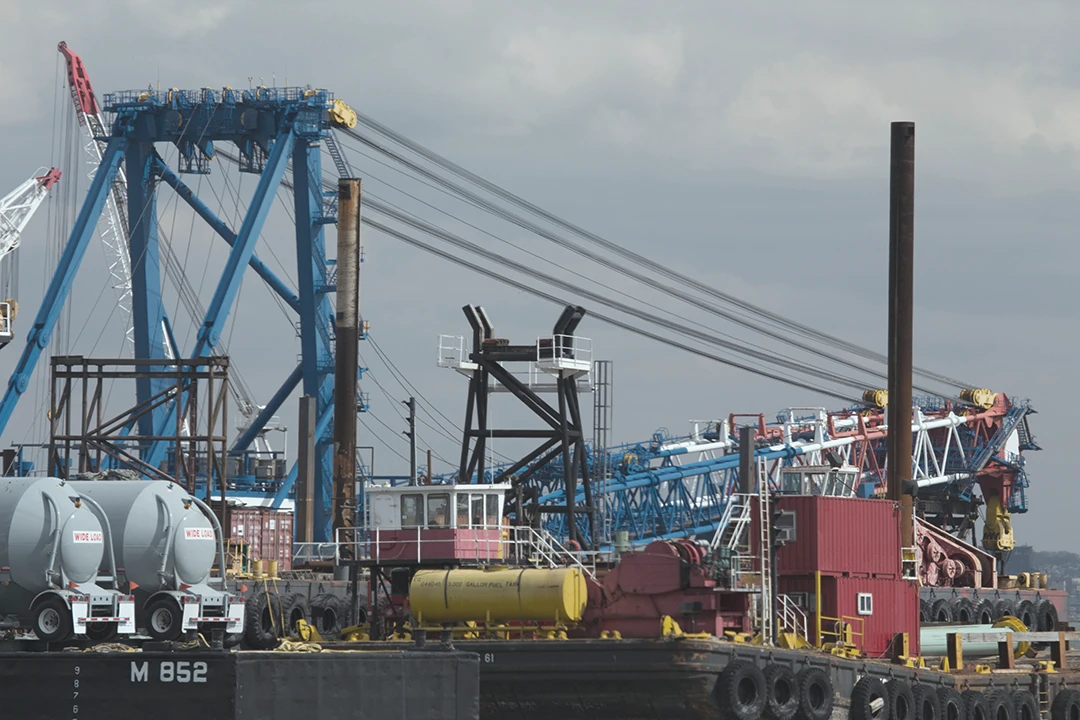 Large cranes, construction equipment, and tanks are stationed on barges at an industrial dock under a cloudy sky, with cables and machinery visible throughout the scene.
