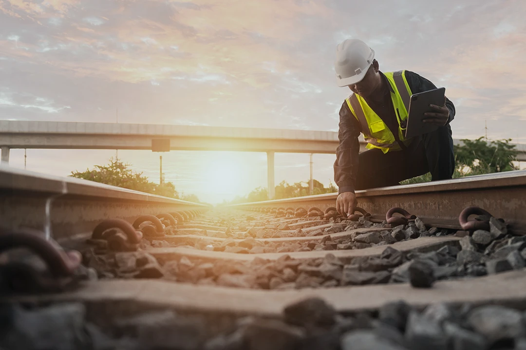 A construction worker in a safety vest and helmet inspects railroad tracks at sunset, holding a tablet and crouching beside the rails. An overpass and sun are visible in the background.
