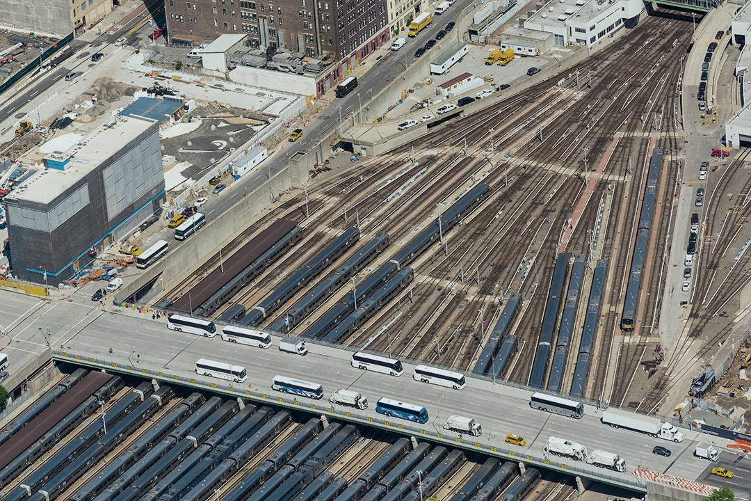 Aerial view of a busy train yard with multiple tracks and trains. A bridge with several buses crosses over the tracks. Buildings and construction sites are visible nearby.