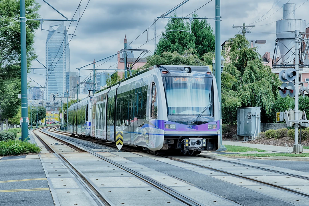A modern light rail train travels through a city, surrounded by greenery and tall buildings under a cloudy sky. Tracks and power lines are visible in the foreground.