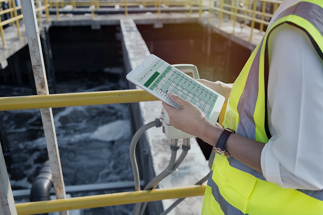 A person wearing a yellow safety vest holds a tablet displaying a spreadsheet, standing near railings at an industrial water treatment facility.