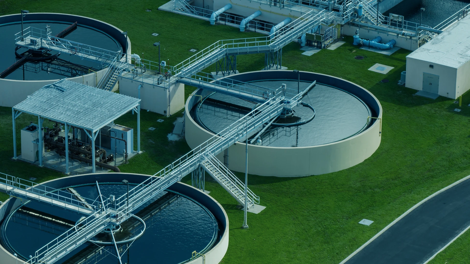 Aerial view of circular water treatment tanks connected by metal walkways at a wastewater treatment facility, surrounded by green grass and paved paths.