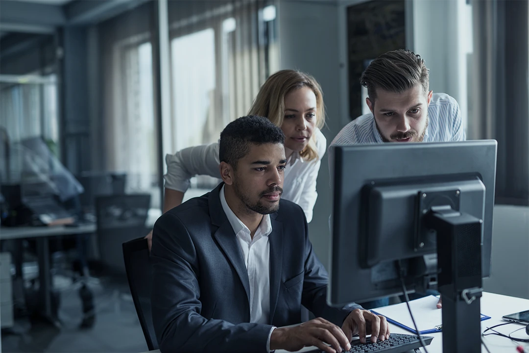 Three colleagues in business attire focus on a computer screen in a modern office, collaborating closely around a desk with office equipment and large windows in the background.