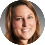 A woman with long brown hair smiles at the camera. The image is a close-up portrait with a gray background and appears slightly blurred.