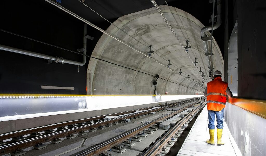 A person in an orange vest and yellow boots stands on the platform of a modern, well-lit train tunnel, facing away from the camera. The tunnel is empty, with tracks running through it.