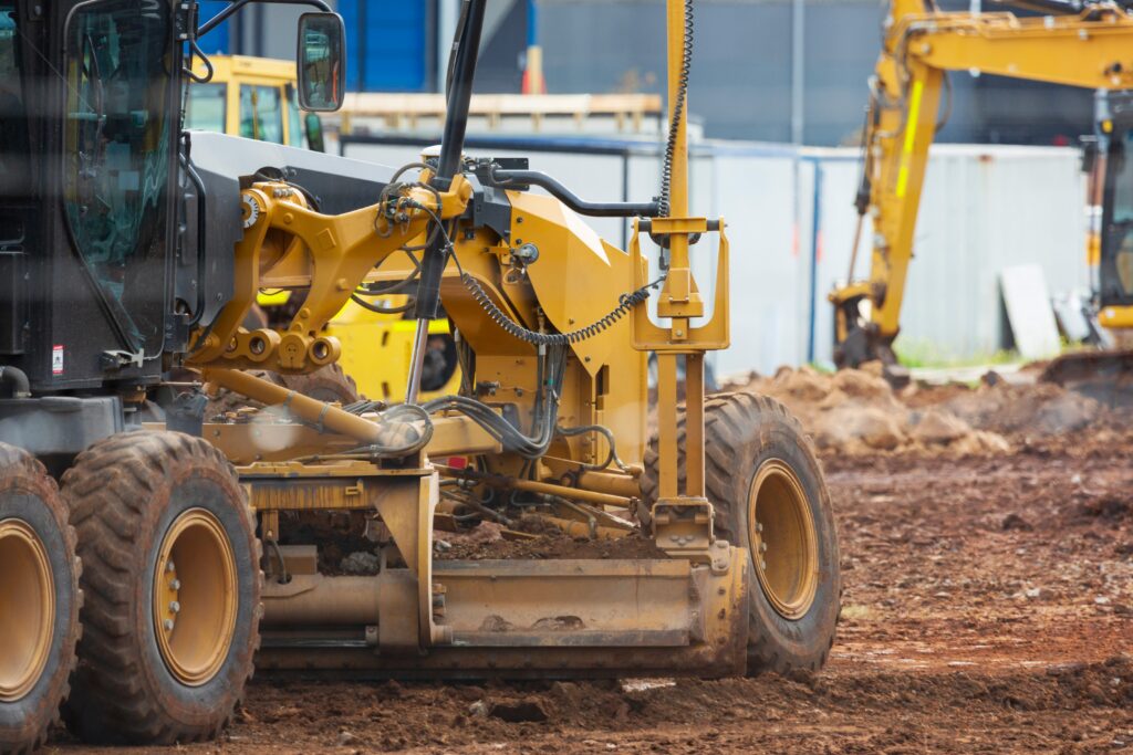 Close-up of a yellow grader working on a dirt construction site, with another excavator and building materials visible in the blurred background.