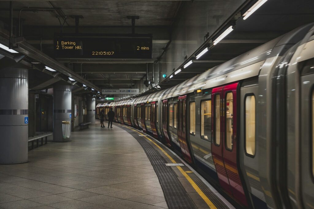A subway train stopped at an underground station; digital sign displays train times. Two people walk on the platform under fluorescent lighting. The environment appears clean and modern.