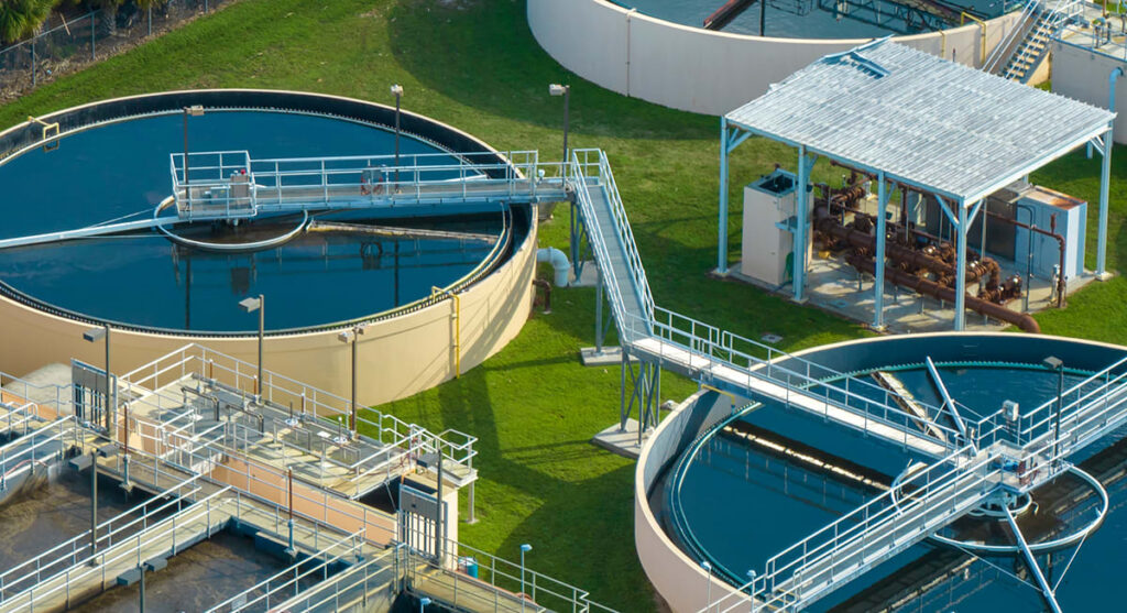 Aerial view of a water treatment plant showing large circular sedimentation tanks and metal walkways surrounded by green grass.