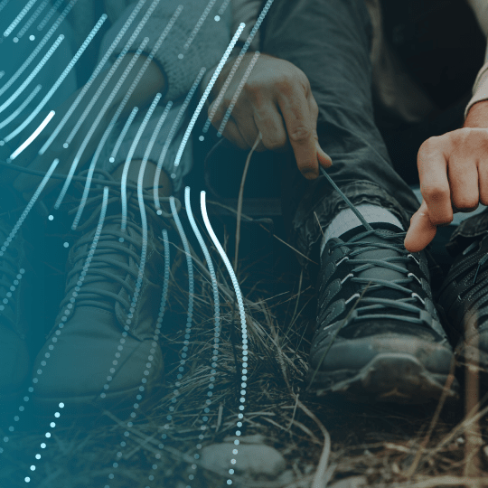 Two people sit on the ground outdoors, tying the laces of their boots. Digital blue lines and dots overlay the image, adding a modern, technological effect.