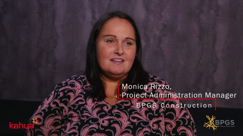 A woman with long dark hair sits in front of a purple backdrop, wearing a black and pink patterned blouse. Text on the image reads: Monica Rizzo, Project Administration Manager, BPGS Construction.