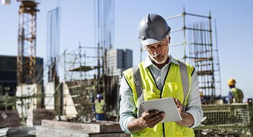 A construction worker in a safety vest and helmet uses a tablet at a building site, with scaffolding, cranes, and other workers visible in the background.