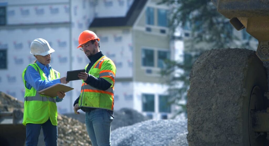Two construction workers wearing safety vests and helmets review documents and a tablet at a building site with gravel, construction equipment, and a partially built house in the background.