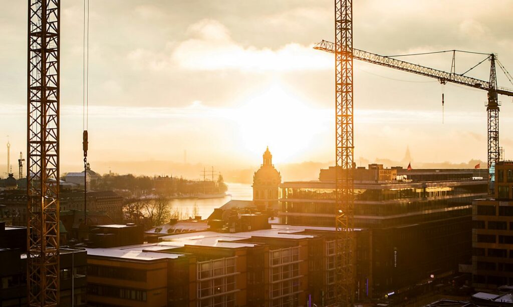 Sunset over a cityscape with construction cranes, buildings, and a river reflecting the sunlight in the background.
