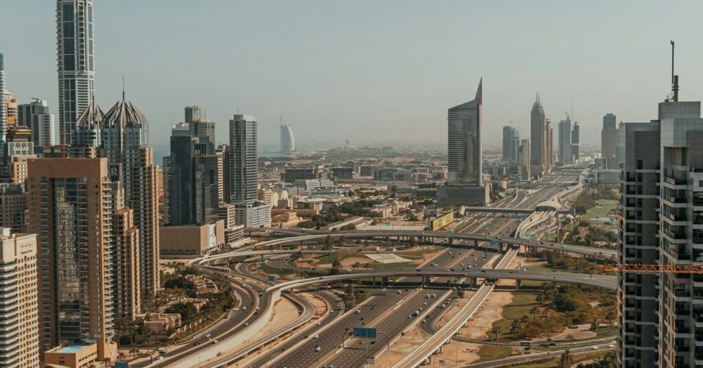 Aerial view of a modern city skyline with skyscrapers, highways, and interchanges under a clear sky. The Burj Al Arab is visible in the distance.