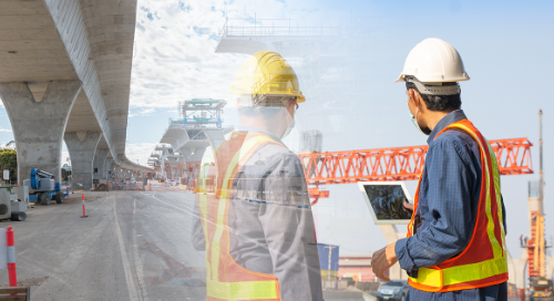 Two construction workers in safety vests and helmets stand on a road next to a large bridge under construction, with cranes and equipment visible in the background.