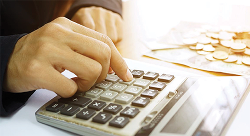 A person presses buttons on a calculator, with coins and paper currency blurred in the background on a bright surface.