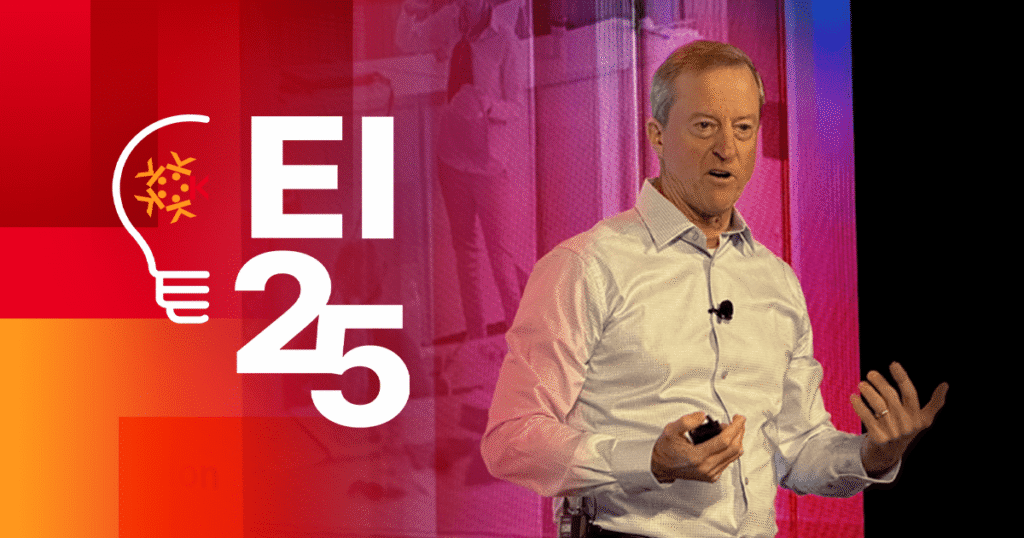 A man in a light shirt speaks on stage, gesturing with one hand and holding a remote. Next to him is a lightbulb graphic and the text “EI 25” on a colorful red and orange background.