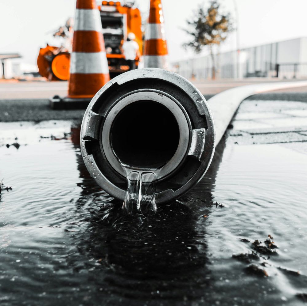 Close-up of water flowing from a large fire hose lying on wet pavement, with orange traffic cones, construction equipment, and a tree visible in the blurred background.