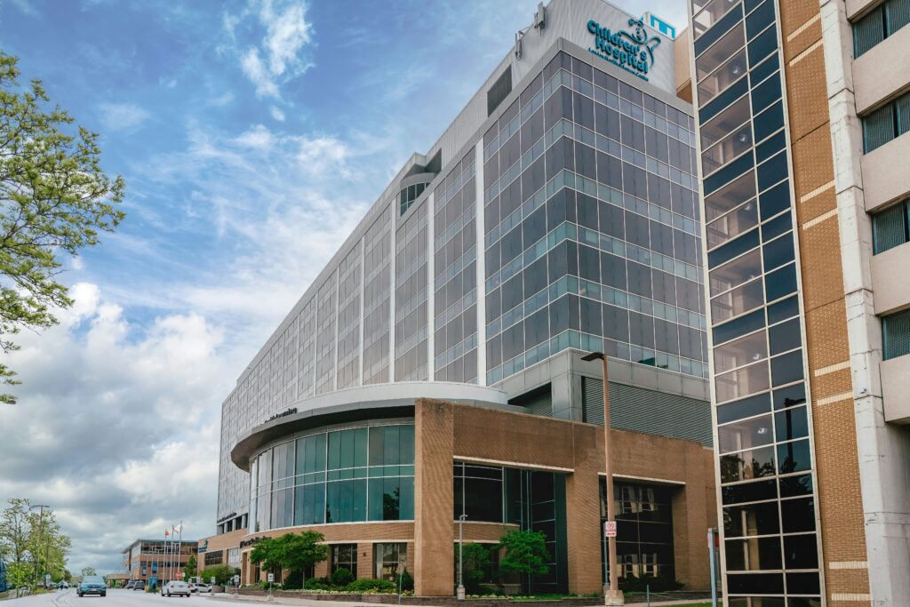 A modern hospital building with large glass windows and a Children’s Hospital logo, adjacent to a street with cars and trees, under a partly cloudy sky.