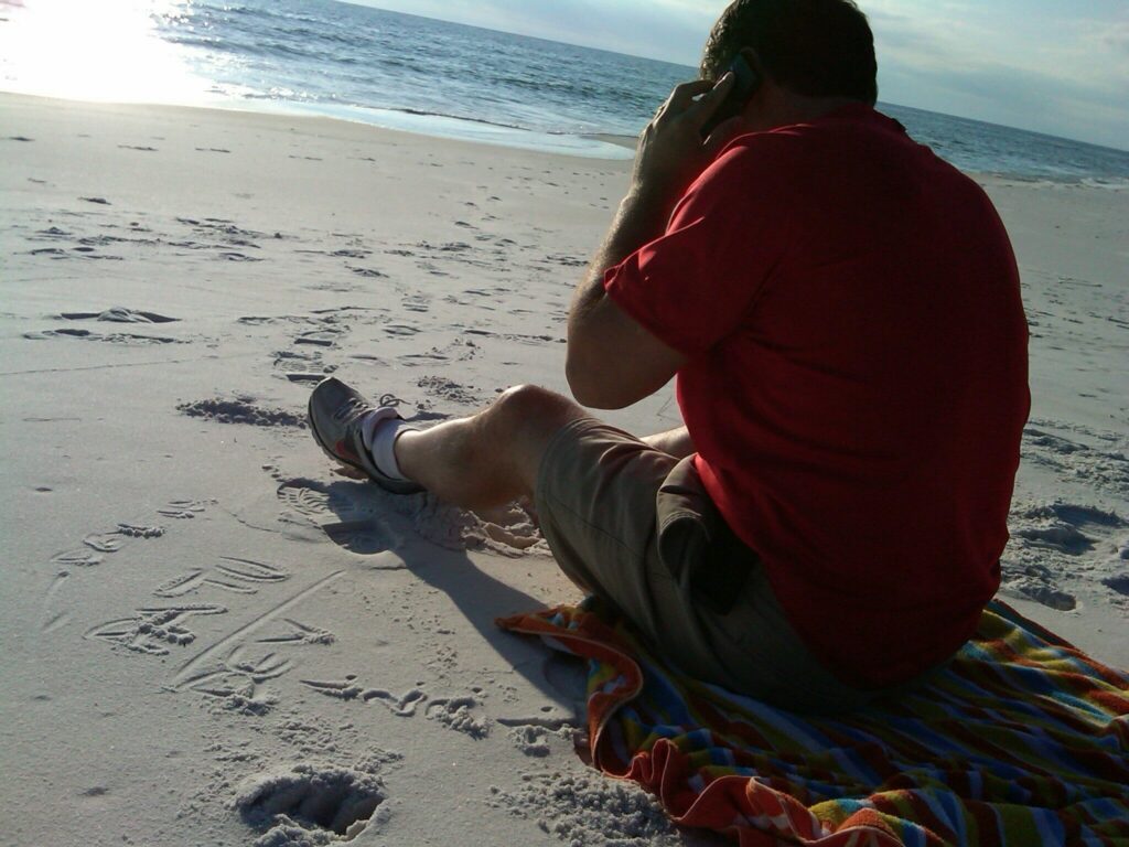 A person in a red shirt sits on a colorful towel at the beach, talking on a phone. The ocean is in the background, and “Beth” with a heart is written in the sand nearby.