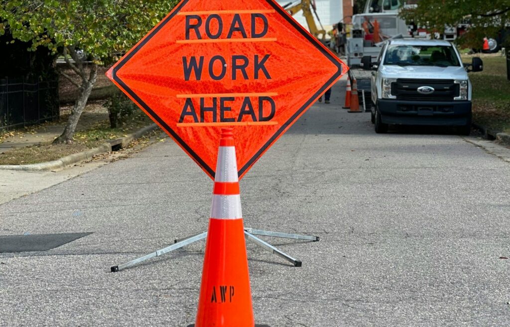Orange Road Work Ahead sign and cone block a residential street; a white utility truck and workers are visible in the background with trees and houses along the roadside.