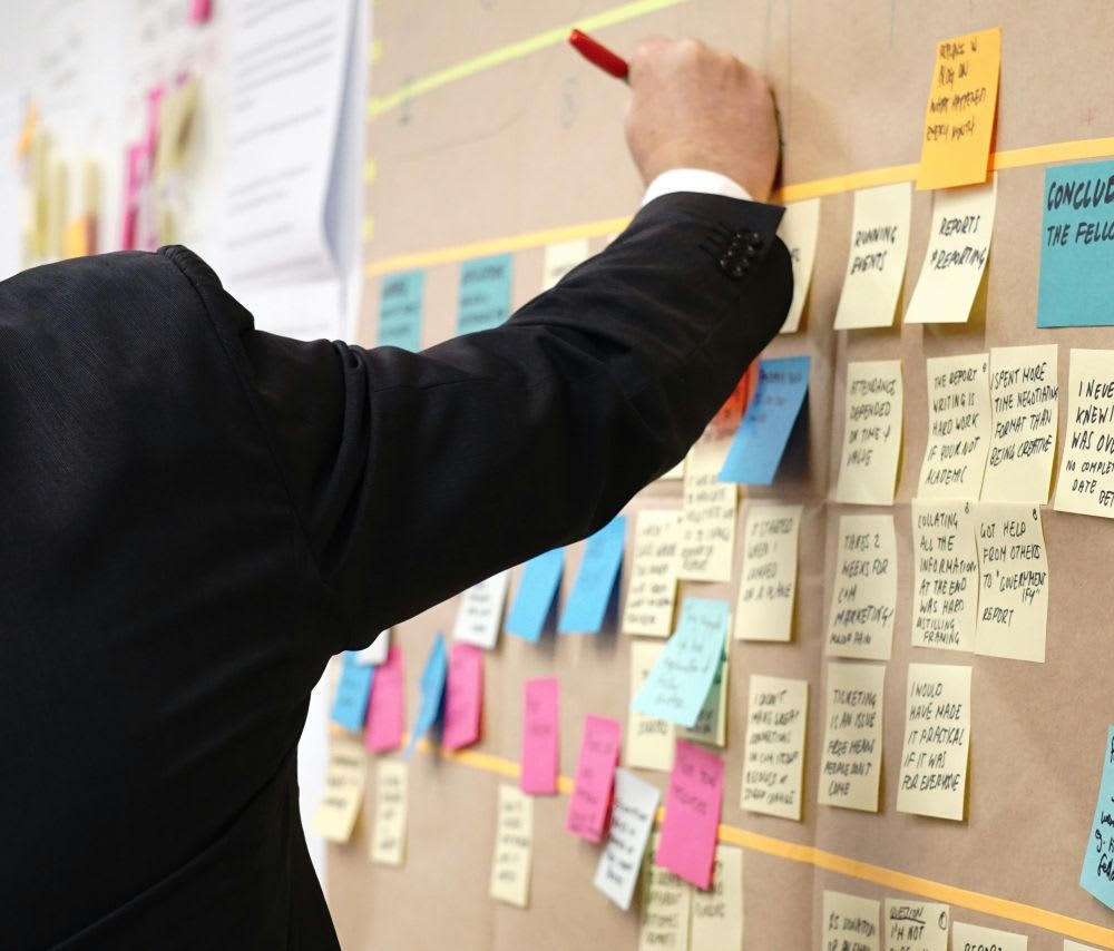A person in a suit writes on a large board covered with colorful sticky notes and documents, suggesting a brainstorming or planning session.