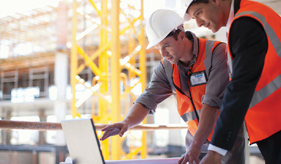 Two construction workers in safety gear look at a laptop on a construction site, with building scaffolding and beams visible in the background.