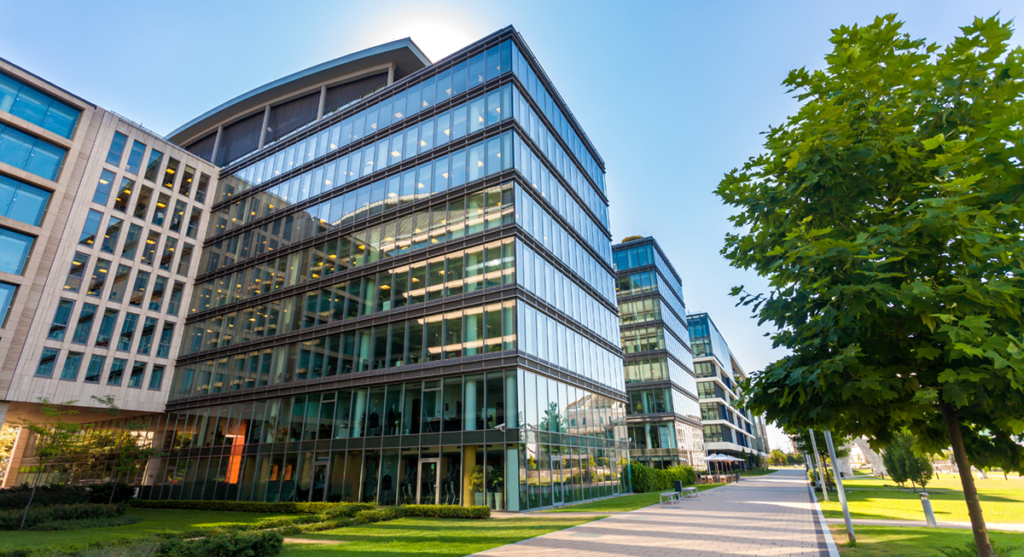 Modern glass office buildings reflect sunlight, surrounded by green lawns and trees, with a sidewalk running alongside. The sky is clear and blue.