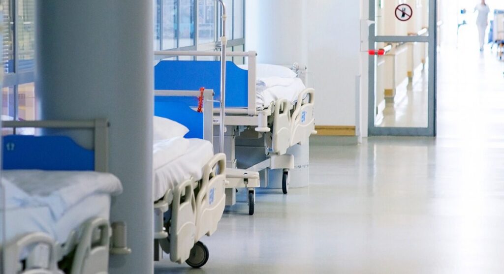 Several empty hospital beds with blue headboards line a clean, brightly lit hallway. The floor is shiny, and a glass door with a no-entry sign is visible at the end of the corridor.