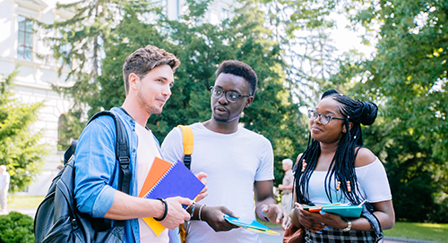 Three students stand outdoors on a sunny day, holding notebooks and backpacks, engaged in conversation in front of trees and a building.