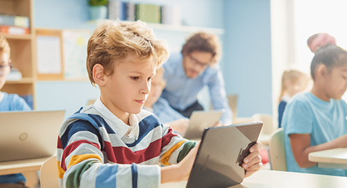 A boy in a colorful striped shirt uses a tablet in a bright classroom with other students and a teacher in the background.