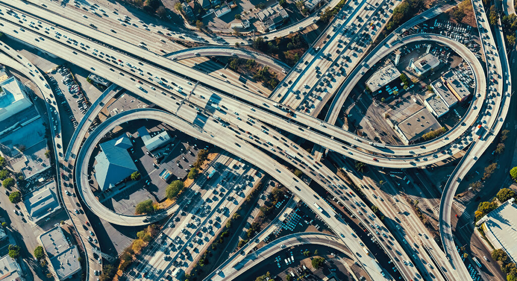 Aerial view of a busy highway interchange with multiple overpasses, looping ramps, and heavy traffic, surrounded by buildings and parking lots.
